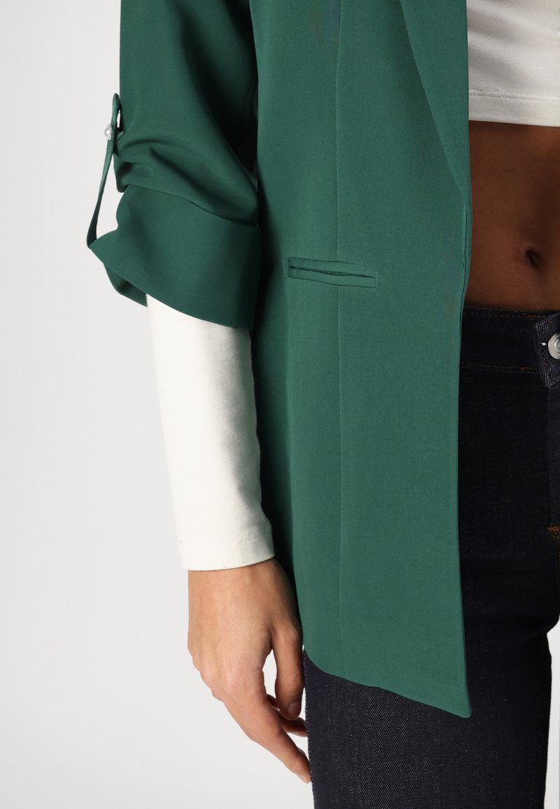 Blazer vert avec des manches retroussées, présentant un léger éclat et une poche avant unique. Assorti à un haut à manches longues blanc et à un jean foncé.