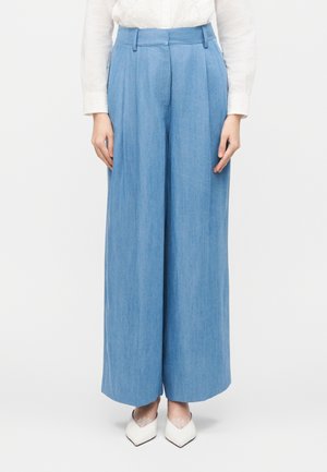 Femme portant un pantalon bleu clair taille haute à jambes larges, un chemisier en dentelle blanche et des chaussures blanches à bouts pointus, sur un fond uni.