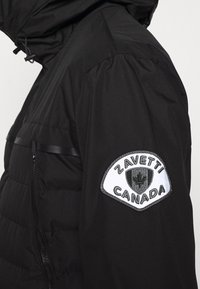 Sort polstret jakkeærme med et hvidt og sort Zavetti Canada-logo patch, der viser et ahornbladsskjold.