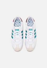 Zapatillas blancas de Adidas con rayas turquesas, punteras beige, lengüetas marrones en el talón y el logotipo de Adidas en la lengüeta y las plantillas, vistas desde arriba.