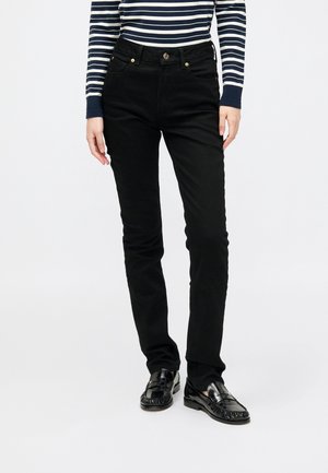 Personne portant un pantalon noir slim, des mocassins noirs brillants et un haut à manches longues rayé bleu marine et blanc, debout devant un fond uni.