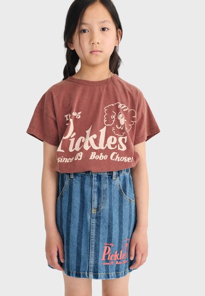Ragazza giovane con lunghi capelli scuri che indossa una maglietta marrone con la scritta "Pickles" e una gonna di denim a righe blu con testo coordinato.
