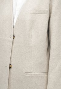 Jasnoszara marynarka w jodełkę z fakturowanej wełnianej tkaniny, z dwoma guzikami i kieszenią na piersi. Prosty, czysty krój. Widoczna biała koszula pod spodem.