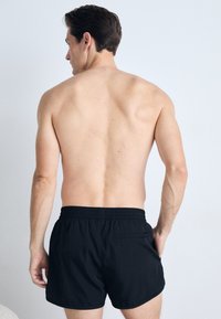 Μαύρο μαγιό με ελαστική μέση και πίσω τσέπη, παρουσιασμένο σε αρσενικό μοντέλο που κοιτάζει μακριά, αναδεικνύοντας την υφή και την κοπή του υφάσματος.