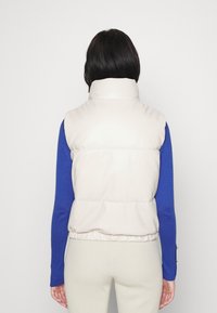 Beige padded vest with a high collar, elastic waistband, and smooth texture, worn over a blue long-sleeve top. Back view.