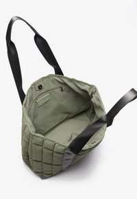 Olivenfarget quilted tote bag med svarte nylonremmer, som har et indre glidelåslomme og glatt stoffinnredning.