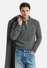 Grauer Fleece-Pullover mit Reißverschluss, kombiniert mit einem hellblau karierten Hemd und blauen Jeans. Auffällige Details sind die gerippten Bündchen und der Kragen.
