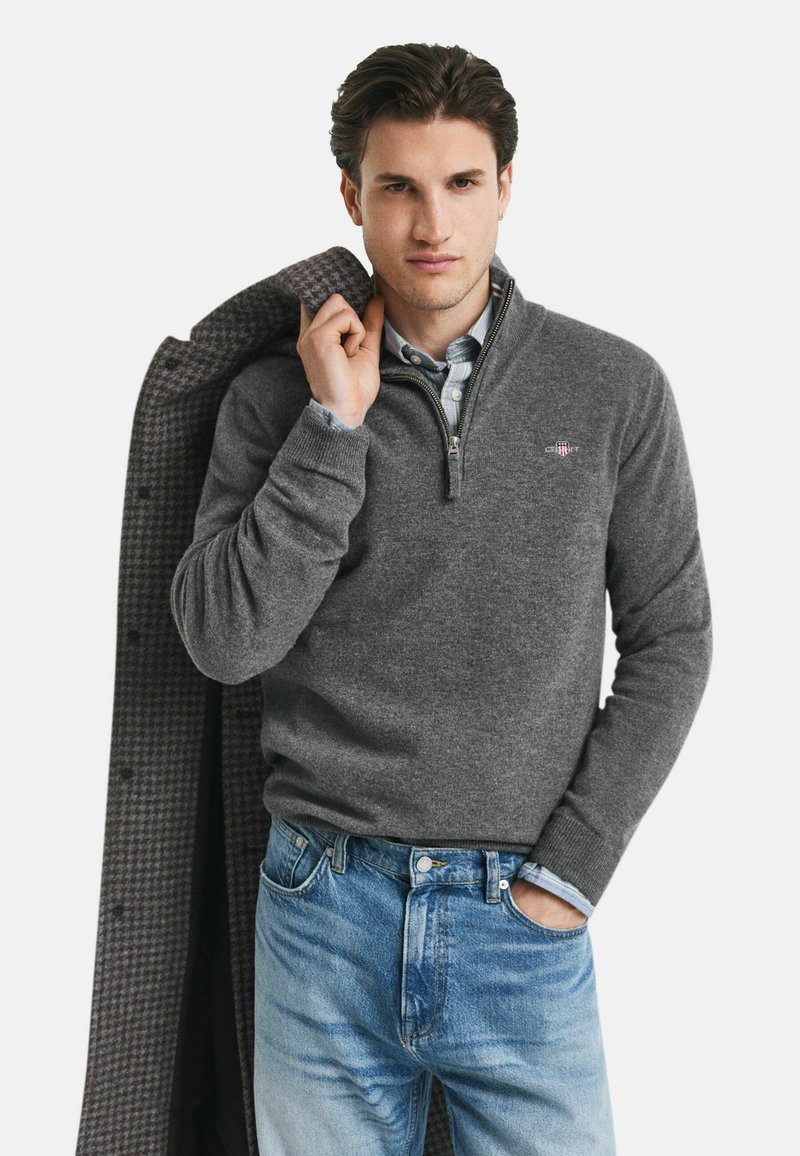 Grauer Fleece-Pullover mit Reißverschluss, kombiniert mit einem hellblau karierten Hemd und blauen Jeans. Auffällige Details sind die gerippten Bündchen und der Kragen.