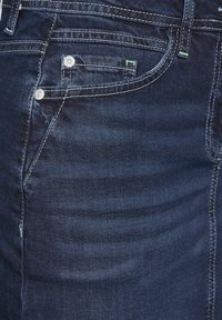 Tmavě modré džíny z denimu s hladkou texturou, které mají přední kapsu, stříbrné kovové knoflíky a kontrastní bílé švy.