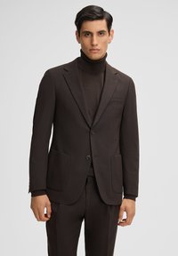 Brauner maßgeschneiderter Anzug mit passendem Rollkragenpullover. Der Anzug verfügt über eine Zwei-Knopf-Verschluss, Reverskragen und zwei Vorderaschen. Strukturiertes Material.