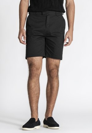 Muž oblečený v čiernych na mieru šitých šortkách a čiernych slip-on topánkach s bielou podrážkou, stojaci na bielom pozadí.