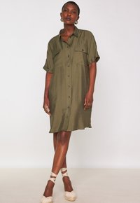 Abito camicia verde oliva con maniche corte, chiusura a bottoni e due tasche sul petto; tessuto leggero con vestibilità comoda. Scarpe espadrille visibili.