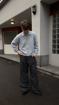 Vaaleanharmaa palmikkoneulepusero, jossa on vetoketjusulku ja kaulus, yhdistettynä väljälahkeisiin tummiin housuihin ja mustiin kenkiin, ulkona märällä pinnalla.