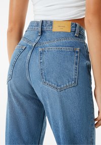 Woman wearing high-waisted blue denim jeans with back pockets and a tan leather patch labeled "Denim Bershka" standing against white background.