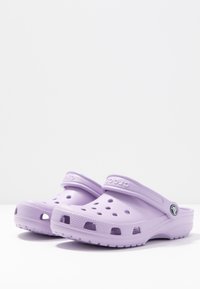 Cizme Crocs violet cu orificii de ventilație, curea la spate și talpă texturată. Fabricate dintr-un material ușor, cu un finisaj neted.