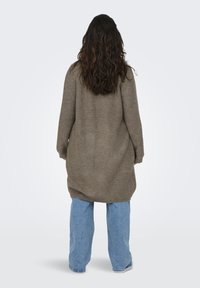 Lång, löst sittande brun cardigan i stickad tyg, med ribbad textur och sänkt axel, kombinerad med blå vida jeans.