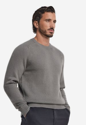 Homme aux cheveux foncés et à la barbe porte un pull en maille gris et un pantalon sombre, debout les mains dans les poches, regardant sur le côté sur un fond uni.