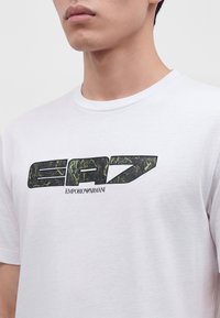 Νεαρό άτομο που φοράει λευκό T-shirt με μεγάλο πράσινο και μαύρο κείμενο "EA7" και μικρότερο λογότυπο "Emporio Armani" κάτω από το κείμενο.