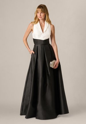 Frau trägt ärmelloses weißes Oberteil und langen schwarzen Rock, hält eine kleine silberne Clutch-Tasche und steht vor einem einfarbigen Hintergrund.