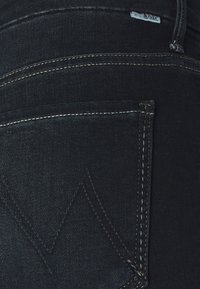 Primer plano de un bolsillo trasero de jeans de mezclilla azul oscuro con costuras en contraste y una pequeña etiqueta de tela de la marca cerca de la cinturilla.