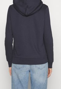 Granatowy hoodie o luźnym kroju, z ribbowanym dołem i mankietami. Posiada kaptur i wykonany jest z miękkiego, teksturowanego materiału. Dobrany z jasnoniebieskimi dżinsami.