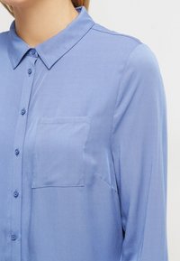 Camisa de botón azul claro con cuello, mangas largas y un bolsillo en el pecho. Textura suave y un brillo sutil.