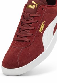 Raudoni zomšiniai sportbačiai su balta guma padanga. Įrengti su auksiniu Puma logotipu ir raišteliu. Textūruota paviršius su matomais siūlių detalėmis.