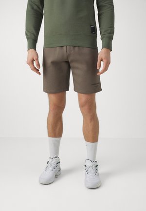 Uomo con felpa verde oliva, pantaloncini marroni Björn Borg, calzini bianchi e scarpe da ginnastica bianche, in piedi davanti a uno sfondo semplice.