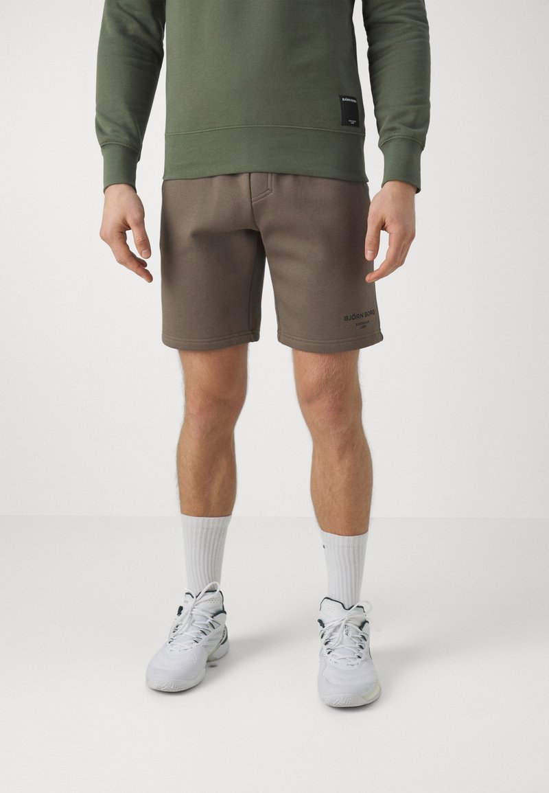 Homme portant un sweat-shirt vert olive, un short Björn Borg marron, des chaussettes blanches et des baskets blanches, debout devant un fond uni.