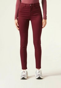 Jeans skinny burdeos con diseño de cinco bolsillos, cierre delantero de cremallera y botón, hechos de tela elástica, combinados con zapatillas deportivas plateadas.