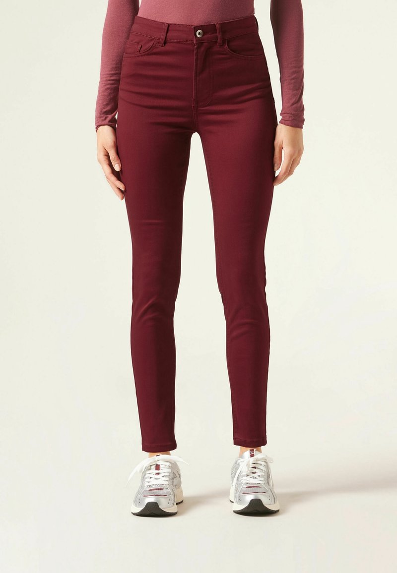 Jeans skinny burdeos con diseño de cinco bolsillos, cierre delantero de cremallera y botón, hechos de tela elástica, combinados con zapatillas deportivas plateadas.