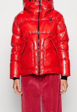 Błyszcząca czerwona kurtka puchowa z wysokim kołnierzem i przednim zamkiem błyskawicznym, z dużymi kieszeniami, noszona z czerwonymi spodniami sztruksowymi i czarnymi rękawami pod spodem.