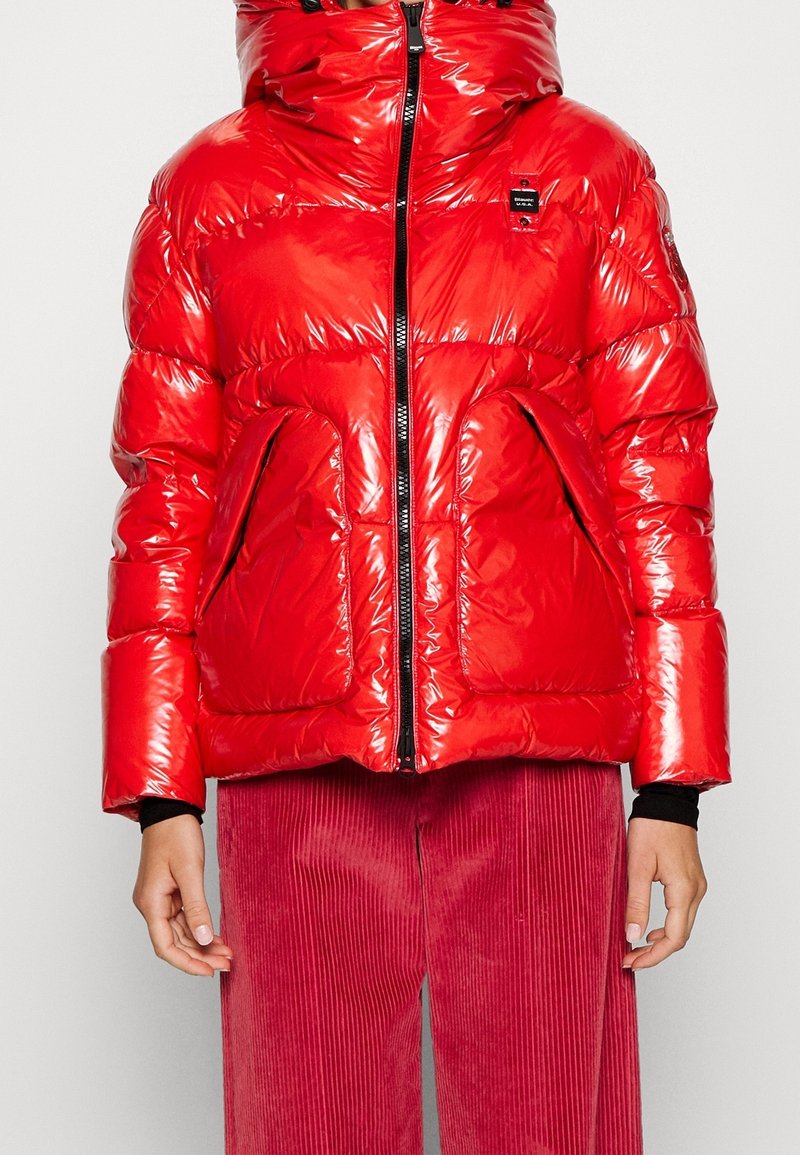 Doudoune rouge brillante avec col montant et fermeture éclair frontale, poches surdimensionnées, portée avec un pantalon en velours côtelé rouge et des manches noires en sous-couche.