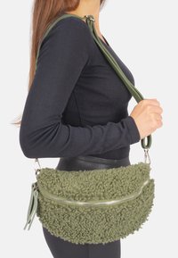 Sac à main vert olive avec un extérieur texturé et en peluche, détail de fermeture éclair argentée et une longue bandoulière adjustable. Dispose d'une forme arrondie.