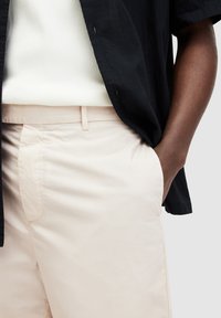 Persona che indossa un top bianco a coste, pantaloni beige e una camicia nera a maniche corte, con una mano in tasca contro uno sfondo semplice.