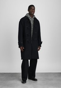 Άνδρας που στέκεται φορώντας ένα μακρύ μαύρο παλτό ντάφλ πάνω από μια μαύρη και λευκή πλεχτή κουκούλα, μαύρο παντελόνι σε φαρδιά γραμμή και μαύρα παπούτσια μπροστά σε γκρίζο φόντο.