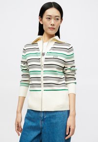 Femeie cu păr negru drept, purtând un pulover alb cu fermoar, cu dungi negre și verzi, și blugi, stând împotriva unui fundal simplu.