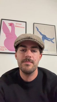 Persona dai capelli scuri e barba che indossa un berretto grigio e una felpa nera. Due manifesti incorniciati con illustrazioni di uccelli sullo sfondo.