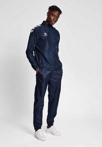 Marinklippad träningsoverall i lätt material med full dragkedja och elastiska ärmslut, försedd med två sidofickor, kompletterad av vita sneakers.