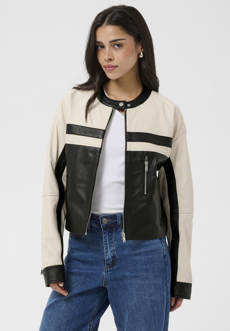 Jeune femme portant une veste en cuir noire et beige sur un top blanc côtelé et un jean bleu, debout devant un fond uni.