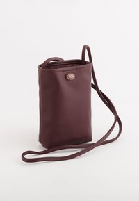 Burgundská kabelka přes rameno vyrobená z texturovaného materiálu s kulatým logem a nastavitelným popruhem, která má minimalistický design.