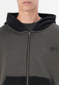 Sweatshirt com fecho zip na cor verde escuro, com capuz preto. Apresenta um fecho na frente, punhos canelados e detalhes nos bolsos. Tecido suave com uma textura misturada.