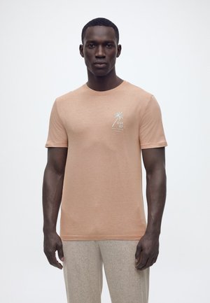 Homme portant un T-shirt marron clair avec un graphisme d'un palmier blanc et du texte sur la poitrine, assorti à un pantalon beige clair.