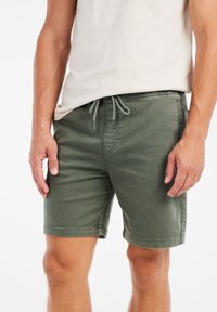 Olijfgroene katoenen shorts met een trekkoordtailleband, voorzien van zijzakken en een relaxte pasvorm, getoond op een model tegen een effen achtergrond.