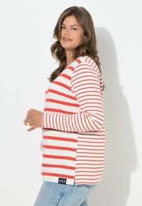 Langärmliges gestreiftes Hemd in Rot und Weiß mit rundem Ausschnitt, Seitenschlitzen und einer Markenetikette am Saum. Blaue Jeans sind sichtbar.