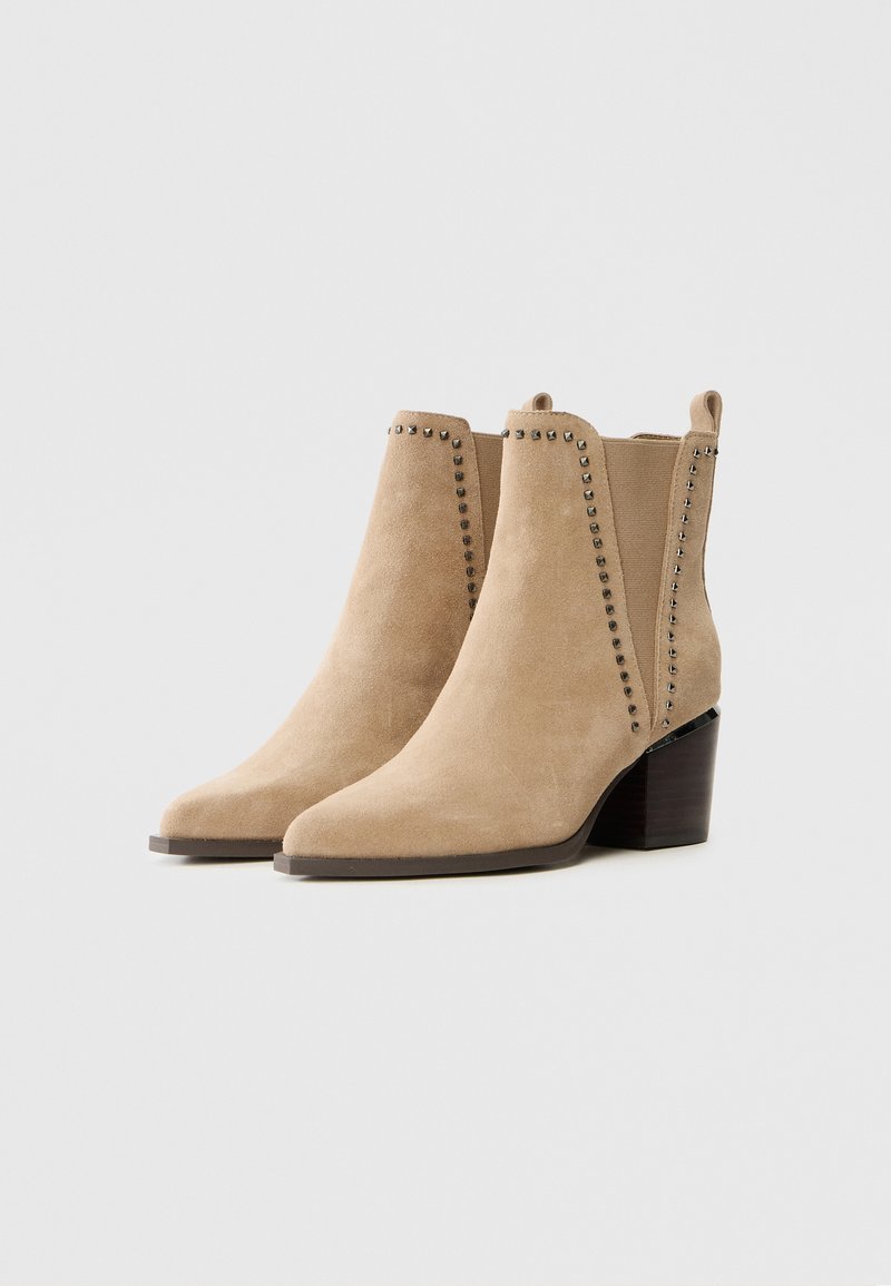 Beżowe zamszowe botki sięgające kostki z wąskim noskiem i grubym obcasem, ozdobione dekoracyjnymi metalowymi ćwiekami wzdłuż szwów bocznych.