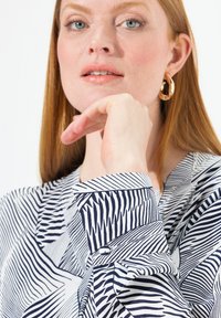 Camicia bianca con motivo geometrico blu navy, con polsini e colletto abbottonati. Il modello ha lunghi capelli rossi e orecchini a cerchio dorati.