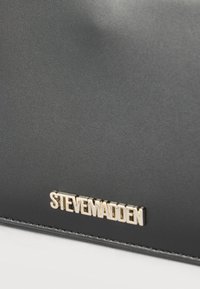 Κοντινή λήψη μαύρης δερμάτινης επιφάνειας με χρυσό λογότυπο της μάρκας "STEVE MADDEN" κοντά στη ραμμένη άκρη.