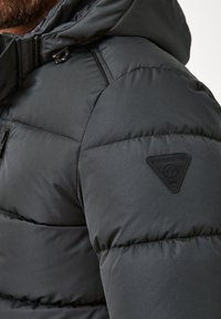 Schwarze Pufferjacke mit strukturierter Oberfläche, ausgestattet mit Kapuze, Reißverschluss vorne und dreieckigem Logo-Patch an der Schulter.