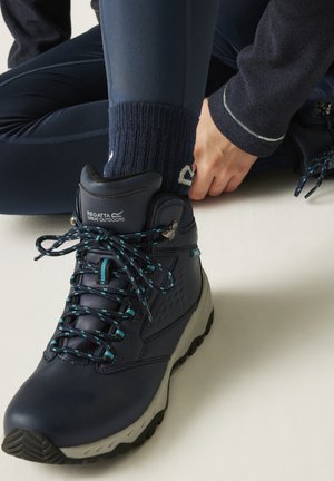 Person tying laces on navy Regatta Great Outdoors hiking boot, wearing dark leggings and navy socks with a white pattern.